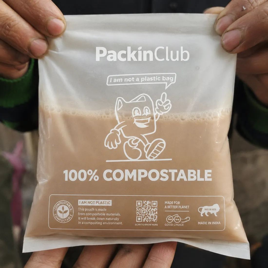 Hands holding a translucent compostable pouch filled with a hot brown liquid, printed 100% compostable